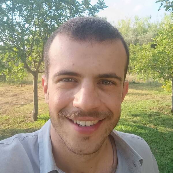 Giuseppe Moledda. A close-up outdoor selfie of a young man smiling warmly. He has short-cropped dark hair, a light mustache, and stubble along his jawline. He is wearing a light grey, open-collared button-up shirt. The background features a sunny, grassy field with green trees under a bright sky.
